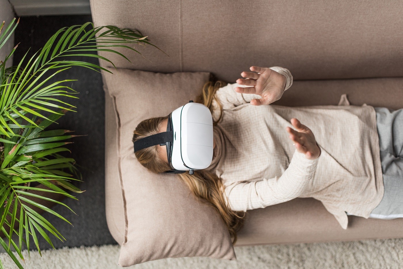 Woman lying on her back on a couch wearing a VR headset. Her hands are in the air as if she is trying to feel something.